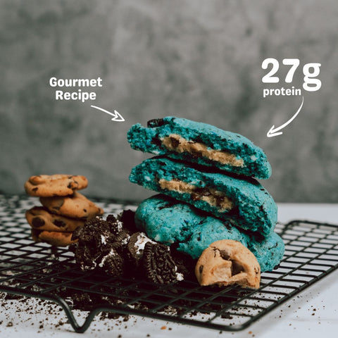 Stack of Cookie Monster cookies and cream protein cookies, featuring vibrant blue color and filled with cookie pieces, with a label highlighting 27g of protein per cookie.